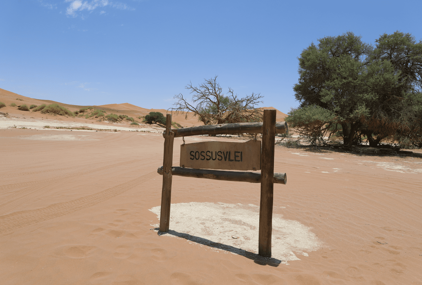 Cartel de madera rústica señalizando la entrada a Sossusvlei en medio del desierto del Namib, rodeado de arena y algunas acacias.